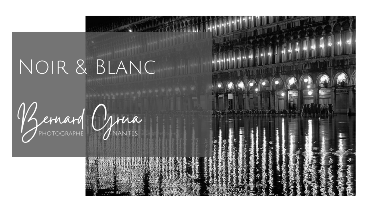Photographie de Bernard Grua en noir et blanc : la place Saint-Marc à Venise illuminée et reflétée dans l’eau.